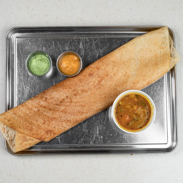 A golden-brown dosa with two small bowls of green and orange chutneys, and a larger bowl of sambar.