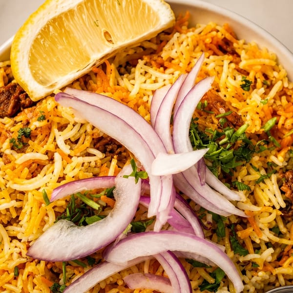 Biryani in a white bowl, garnished with red onions, cilantro, and a lemon wedge.
