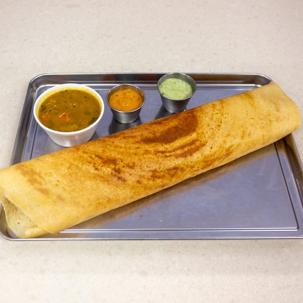 A golden-brown dosa with three small bowls of chutney and sambar on a metal tray.