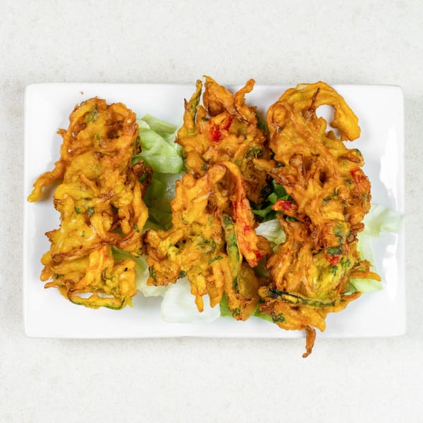 Four golden-brown vegetable pakoras, garnished with lettuce, on a white rectangular plate.