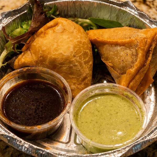 Crispy samosas with tamarind and mint chutneys in a foil takeout container.