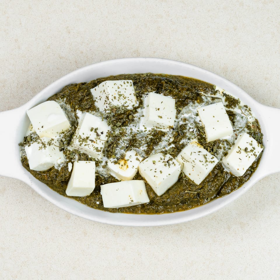 Palak Paneer, a vibrant green spinach curry topped with white paneer cubes and dried herbs in a white dish.
