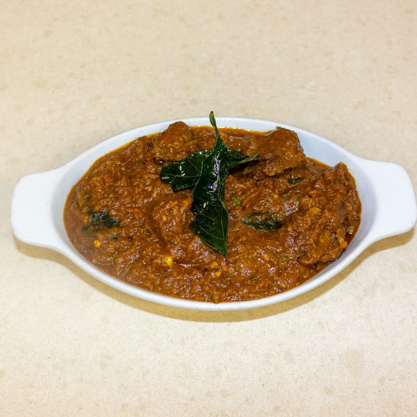 A white oval dish filled with a rich, reddish-brown curry, garnished with fresh green leaves.